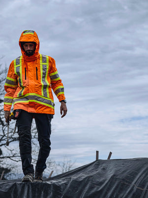 Tradesman wearing an orange safety jacket CSA Hi-Vis on a roof with trees and cloudy sky in the background