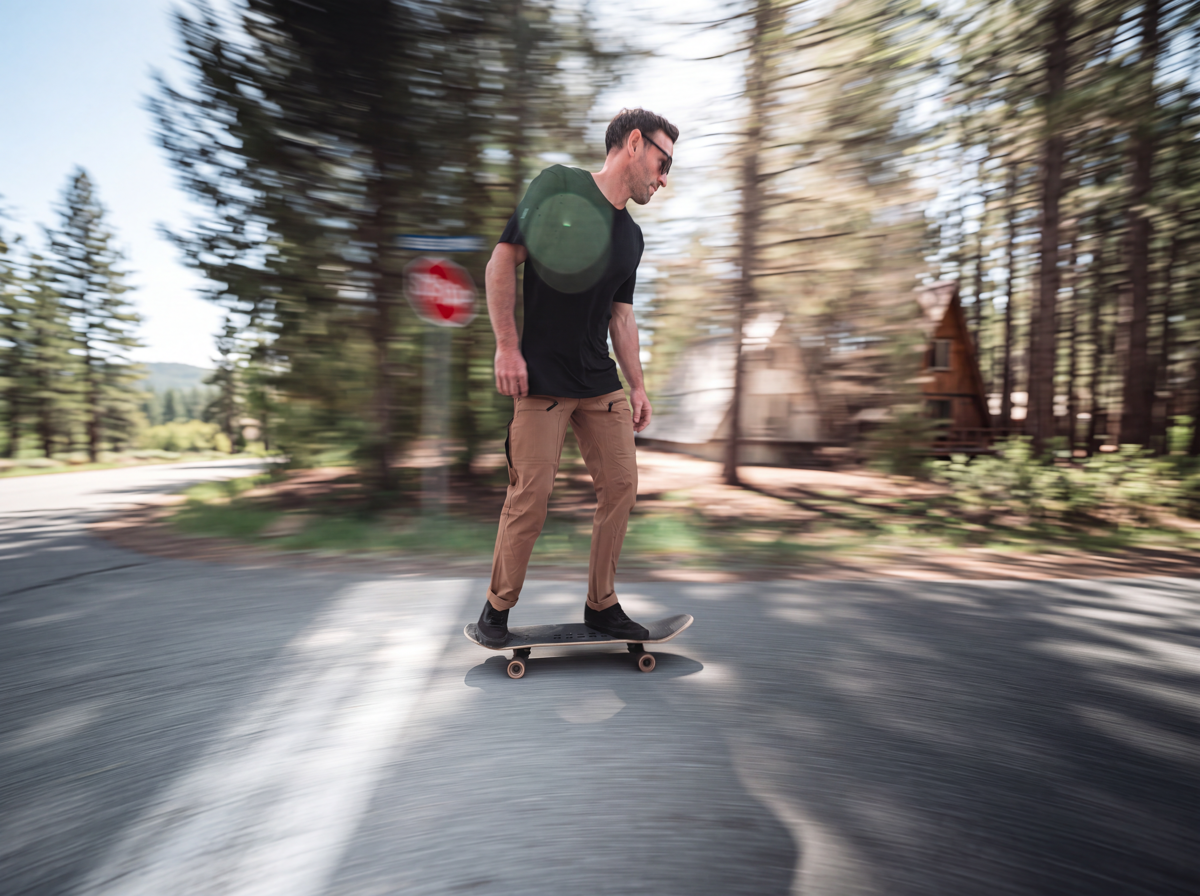 Translation missing: en.A man wearing a black t-shirt, tan pants, and black sneakers rides a skateboard down a paved road surrounded by a blurred forest background. In ventilated blaklader solano pants