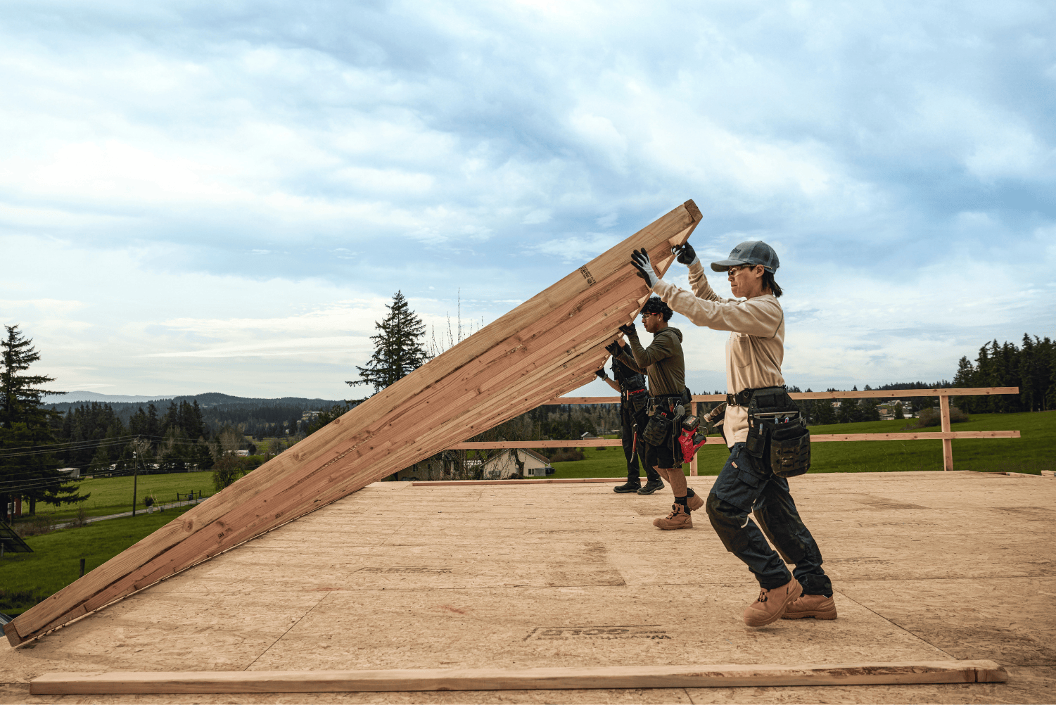 tradeswoman and crew lifting a large wooden panel outdoors with trees and sky in the background